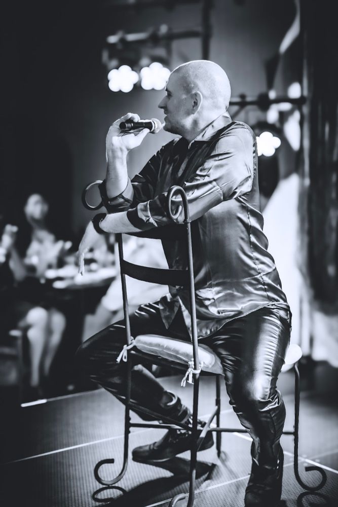 A black and white photo of me sitting backwards in a chair and holding a microphone. The shirt and pants are shiny and reflective in the lighting.