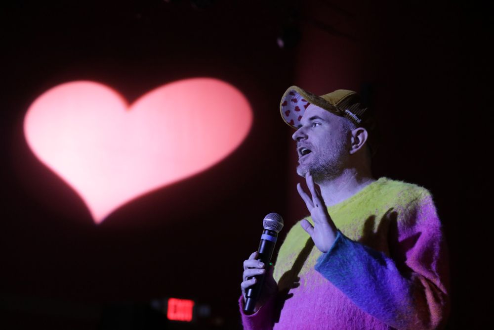 Myself, wearing a loud yellow, pink, purple sweater standing and speaking into a microphone. A light in a heart shape is projected behind me speaking