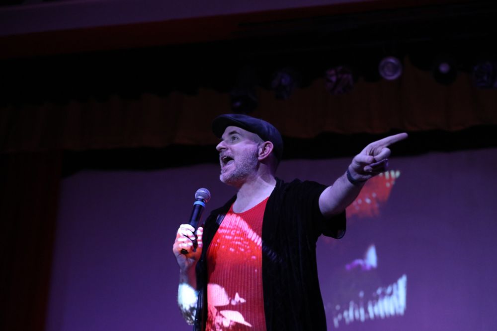 Me, wearing a red shirt and black button up, pointing at something in the audience while the iconic red rocky horror lips are projected on me