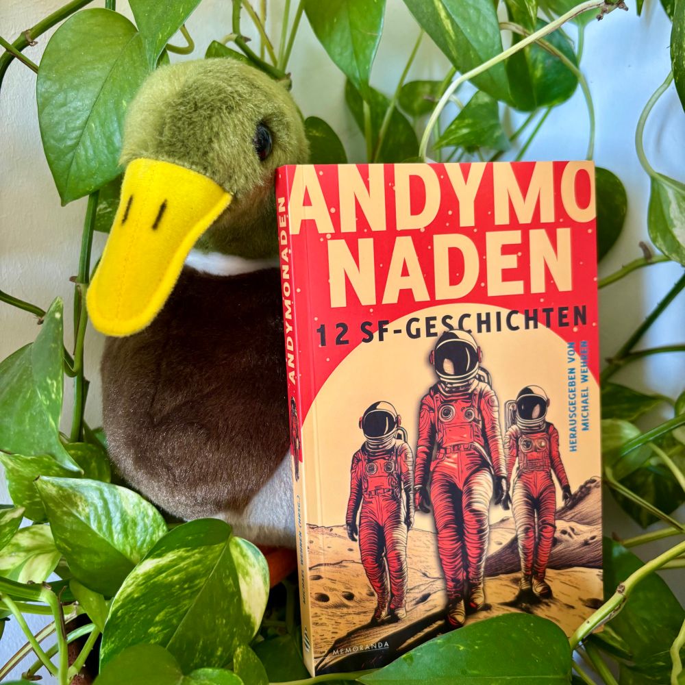 foto der anthologie "andymonaden". auf dem cover sind drei figuren in raumanzügen vor rotem, orangenem und gelben hintergrund. die anthologie lehnt an einer plüschente, weil ein erpel in meiner geschichte vorkommt.