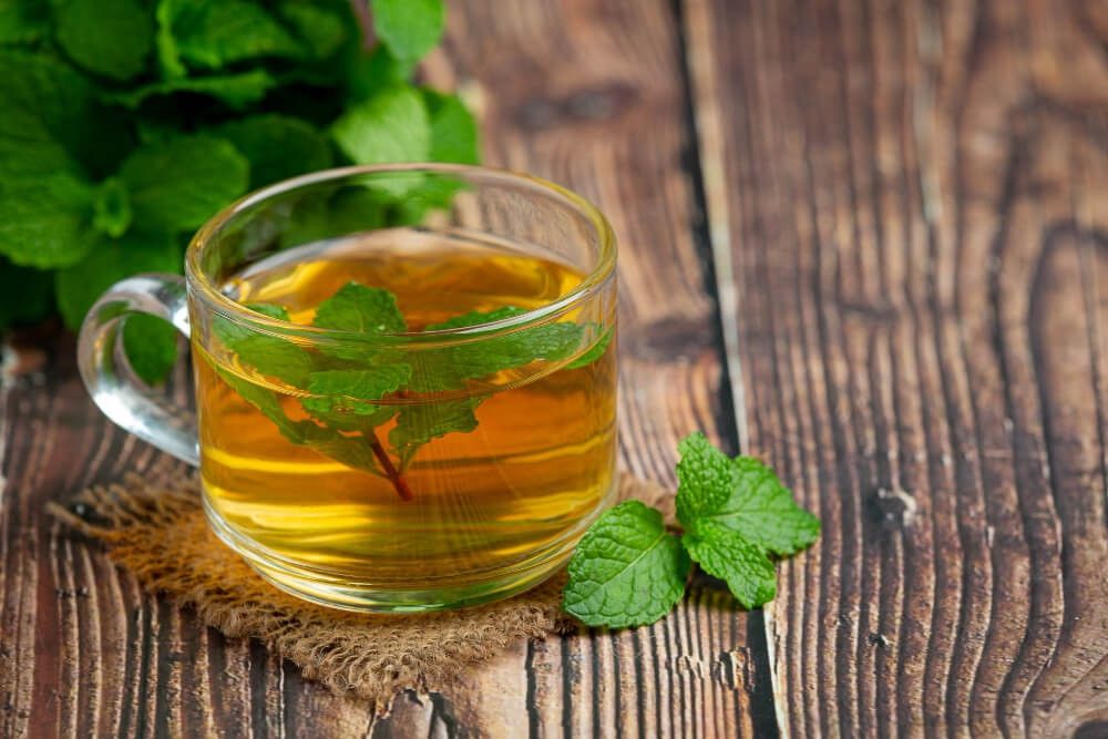 a glass mug of spearmint tea. green spearmint leaves are in the tea, to the side, and big cluster is behind. all sat on a wooden table surface.