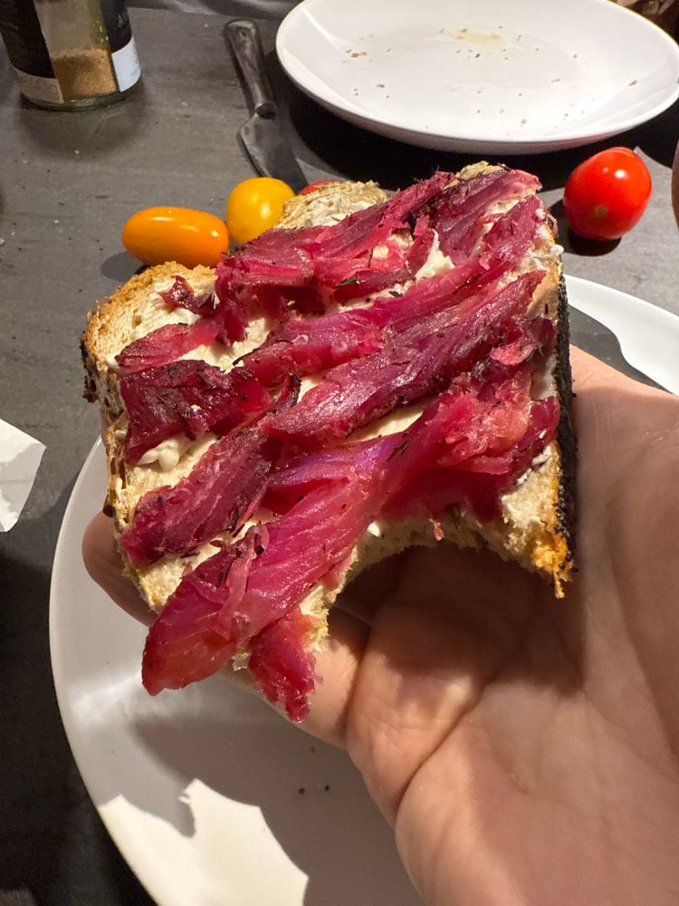 Im Hintergrund ein paar Tomaten, auf meiner Hand ein Weißbrot mit rot gefärbtem Lachs