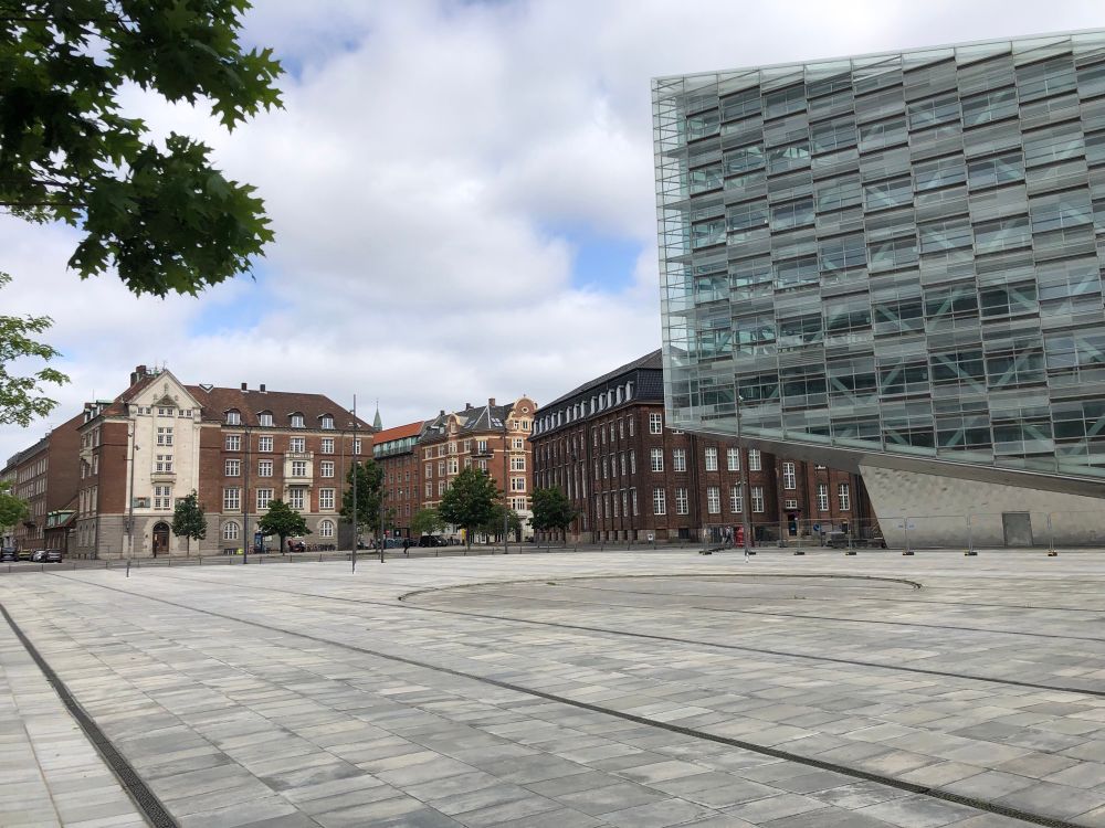 A square with a modern angular building on the right and older brick buildings in the background