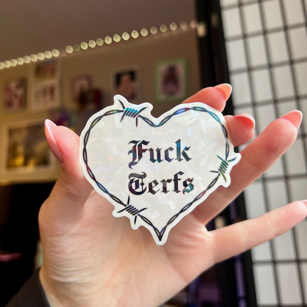 Holographic sticker in the shape of a barbed wire heart that says Fuck Terfs in Old English font