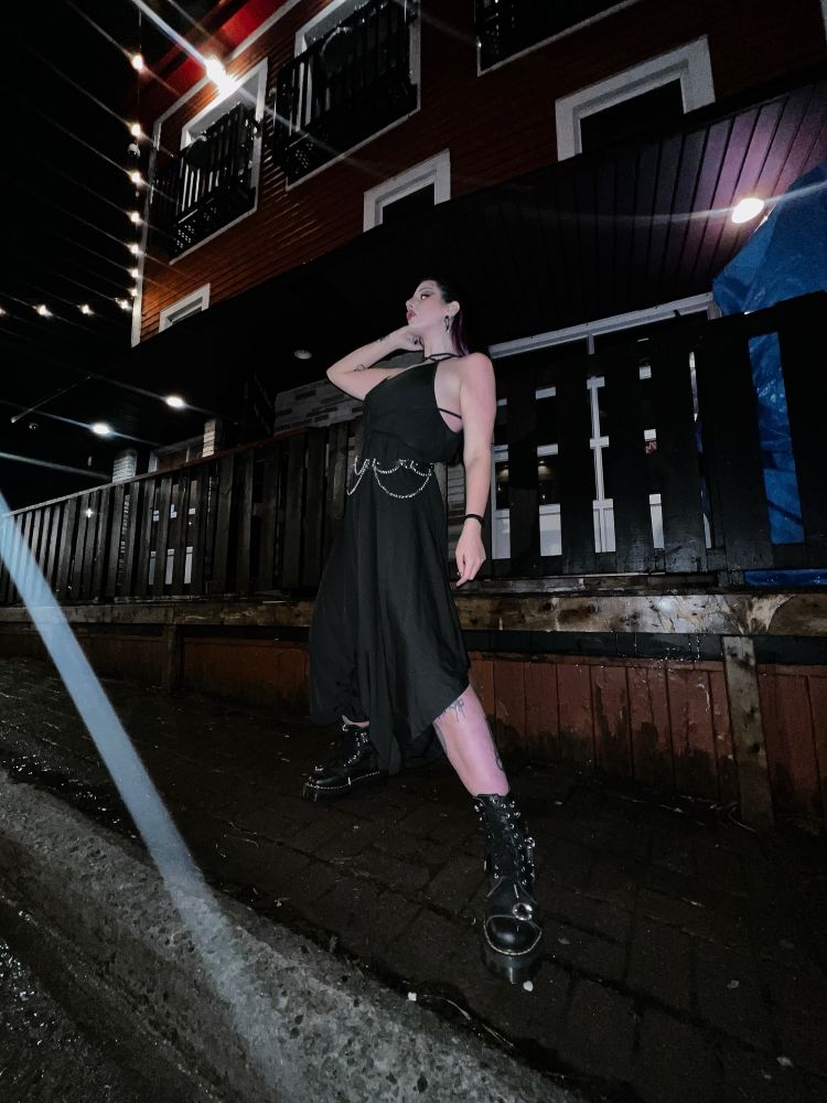 Villanarei wearing a long flowy black jumpsuit with wide legs, a silver chain belt around her waist, and black Dr. Marten boots. Her hair is slicked back, and she’s wearing black heart shaped hoop earrings. 