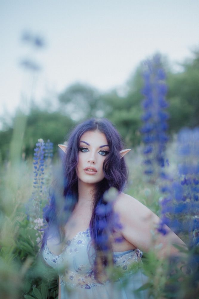 Villanarei sitting in a field of lupins, wearing light blue babydoll lingerie, elf ears, and her purple hair is down and wavy.