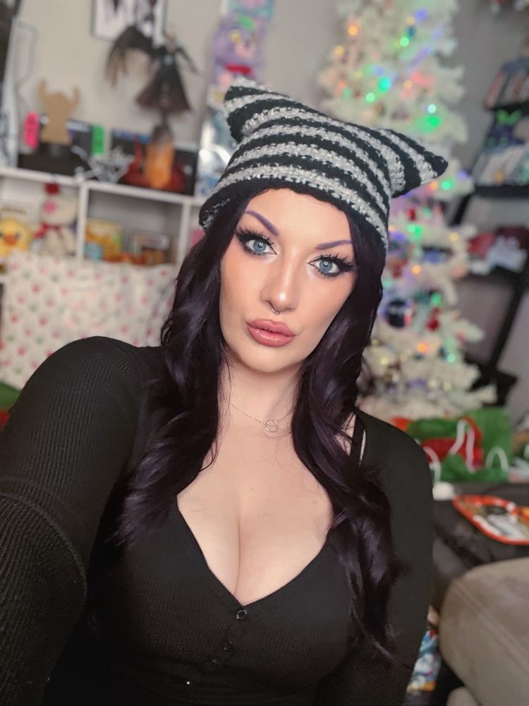 Villanarei wearing a black v-neck long sleeve shirt, and a black and grey striped cat ear hat. Her dark purple hair is curled, and she’s wearing dark eyeliner. 