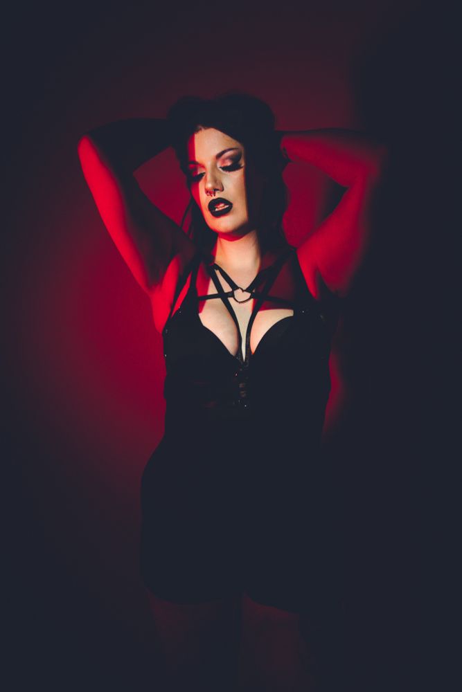 Villanarei wearing a black bodysuit and a black pentagram chest harness. The lighting is red and black, with an inverted cross projected across her face and chest.
