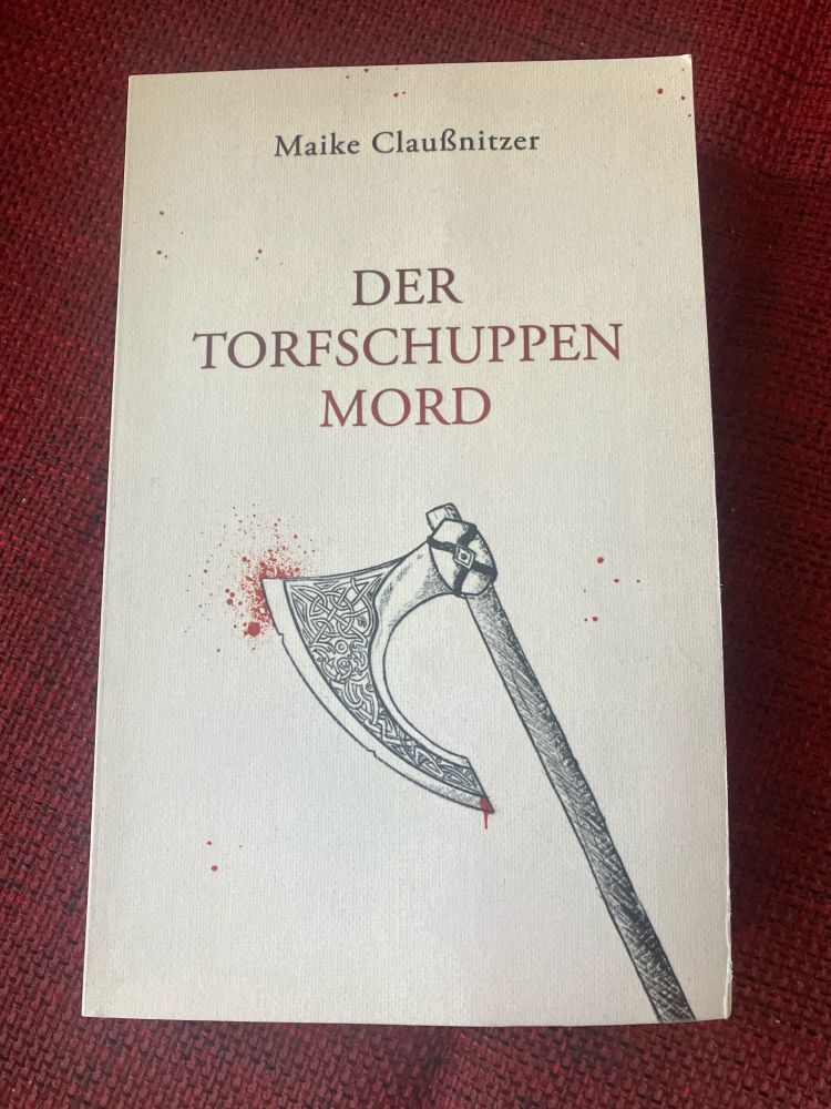 Foto des Buches Der Torfschuppenmord von Maike Claußnitzer
Das Buch hat ein hellgraues Cover, auf dem eine gezeichnete Axt mit keltischem Muster, einige Blutspritzer sowie Name und Titel abgebildet sind. Das Buch liegt auf einem dunkelroten Untergrund.