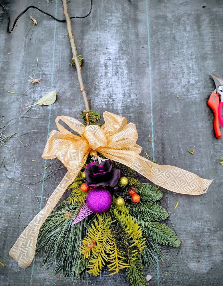 A Witch's Broom made of pine, cedar, and sage, adorned with a glittery purple orb and rose, and several orange and green accents. Tied at the handle with a large gold bow. 