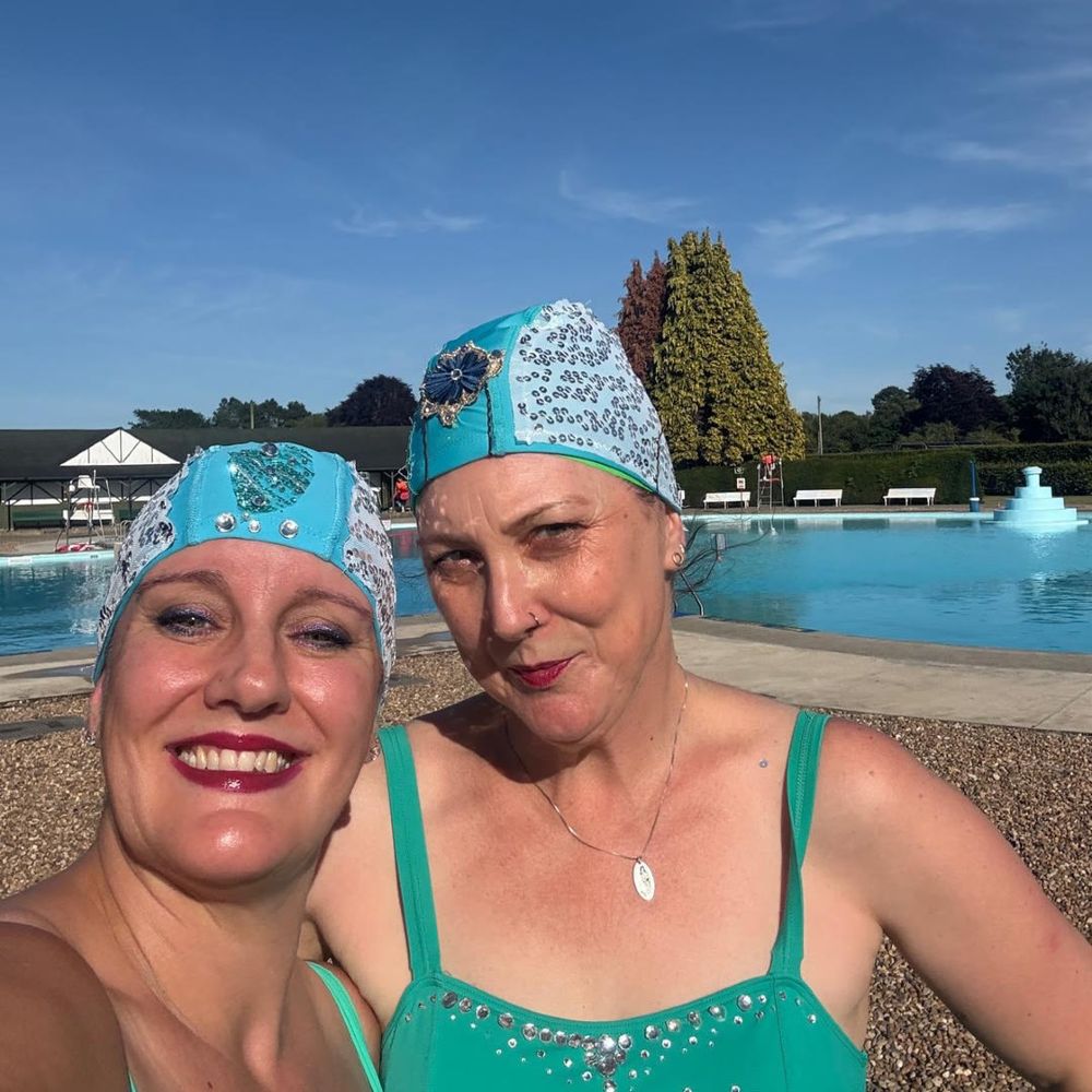 Two women in turquoise swimsuits and embellished swim caps smile at the camera beside an outdoor pool under a clear blue sky.