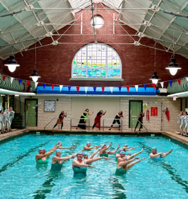 Synchronized swimmers in green caps pose with arms extended in a historic indoor pool, while dancers perform on deck in front of a stained glass window.