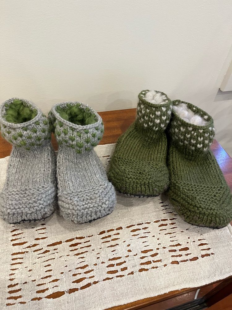 A photo of two pairs of handknit slippers. The pair on the left is knit in gray chunky wool with green thrums around the cuff. The slippers on the right are knit with pine green chunky wool with white thrums around the cuff. A thrum is a strand of unspun wool that is worked into the fabric of a knitted garment to provide cushioning and warmth.