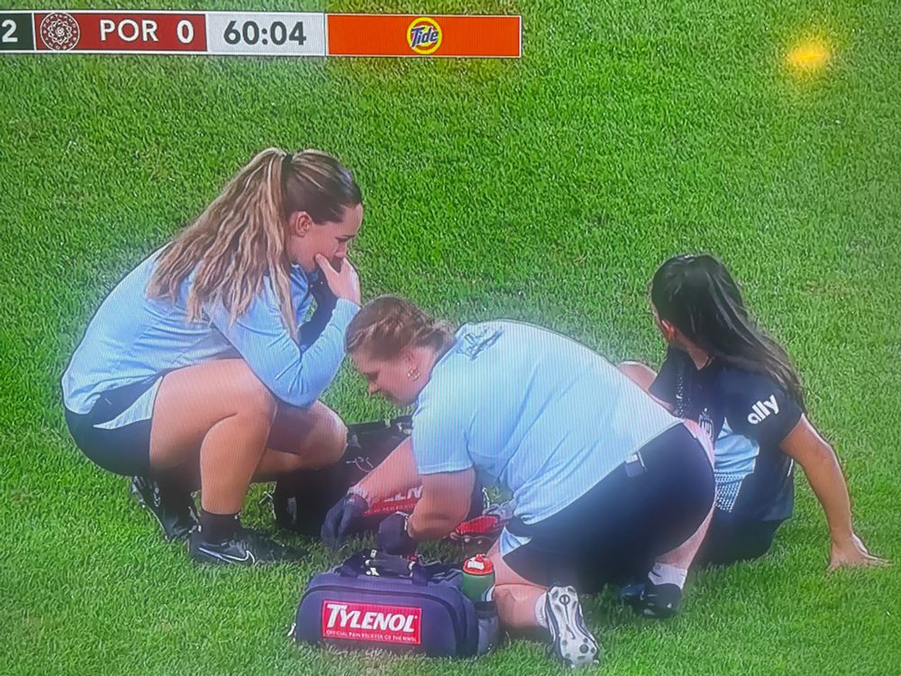 Two Gotham on-field medics attending to an injured player. Their med kit bags have giant Tylenol brand logos on the side, strategically placed directly toward the camera.