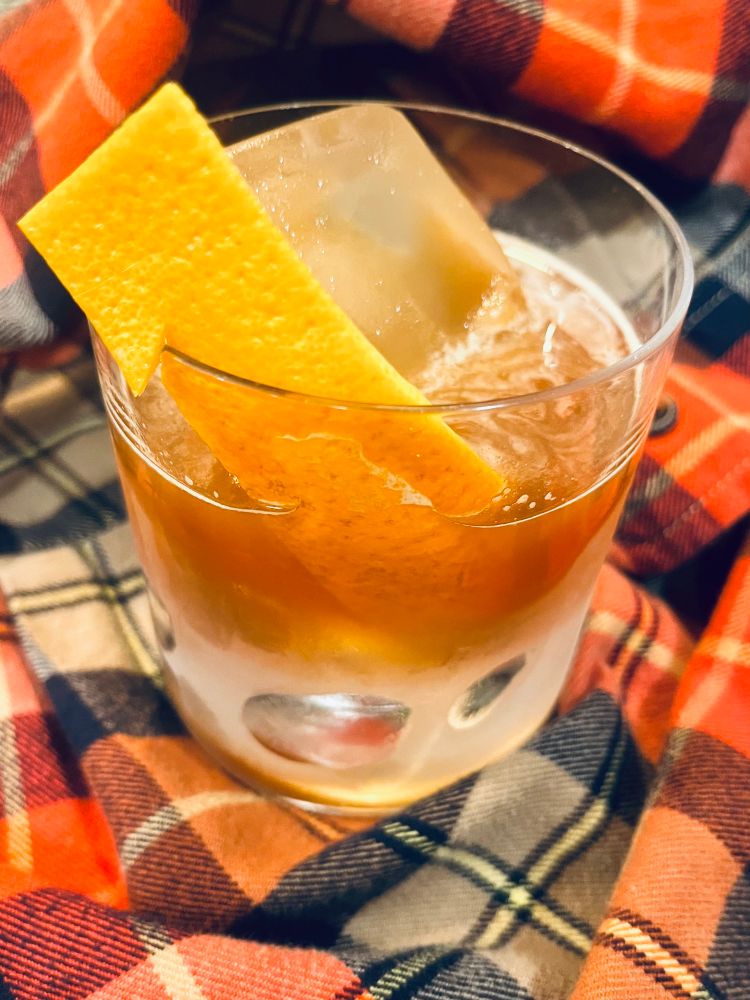 A light brown hazy cocktail in a chilled rocks glass with an orange peel garnish. The glass is sitting on an orange flannel shirt.