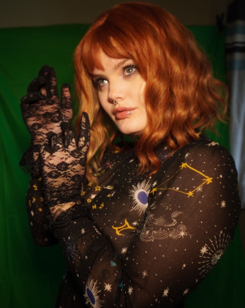 Ezra (they/them) wears a ginger curly bob wig, black floral lace gloves, and a sheer black dress with celestial patterns. Their hands are together near their face and they gaze up into the distance.