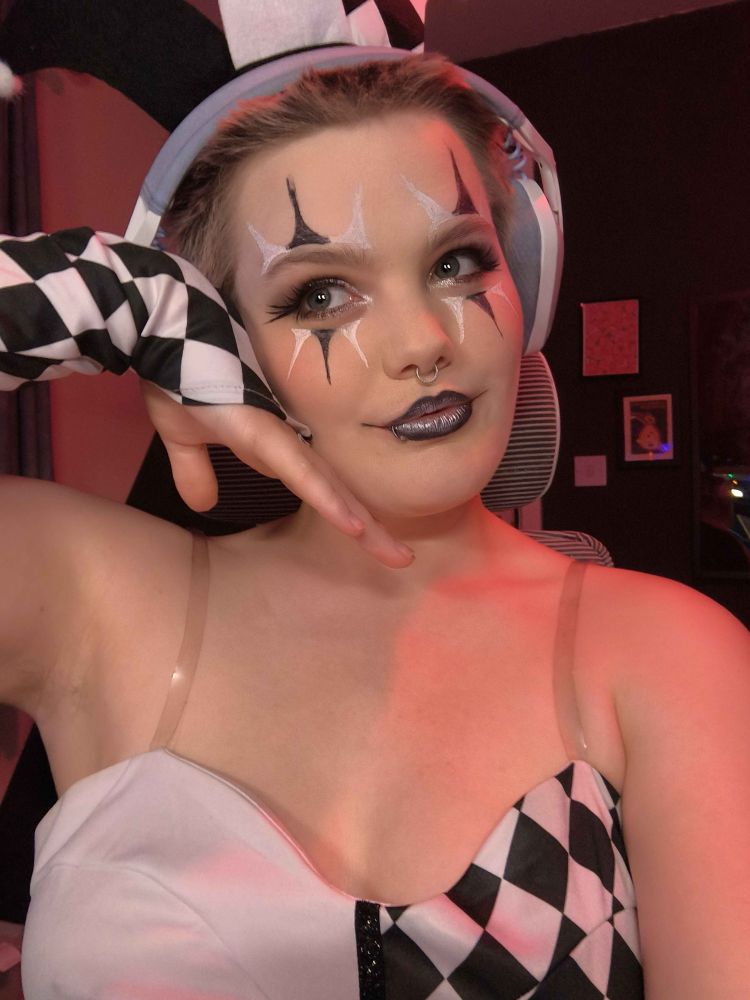 Ezra (they/them) wears a black and white clown dress with black and white clown makeup. They have one hand under their chin and smile demurely, looking to the distance
