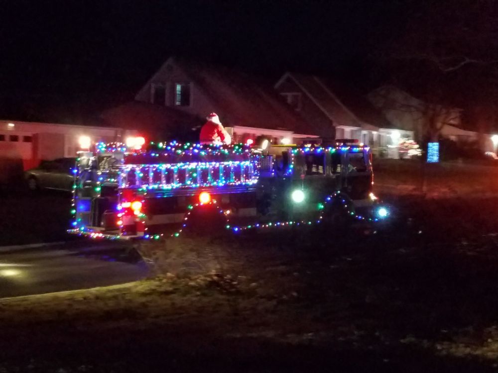 Fire truck lit up with Christmas lights, with Santa on top, driving through a neighborhood.
