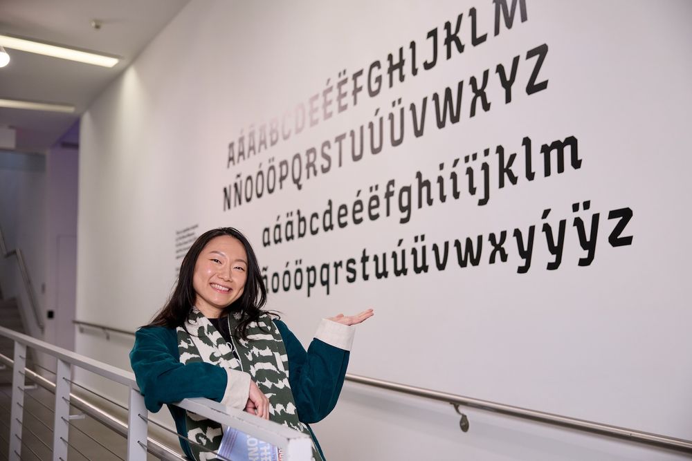 Lynne Yun stands in front of the typeface she created, smiling. The typeface is adhered to the wall, in black. 