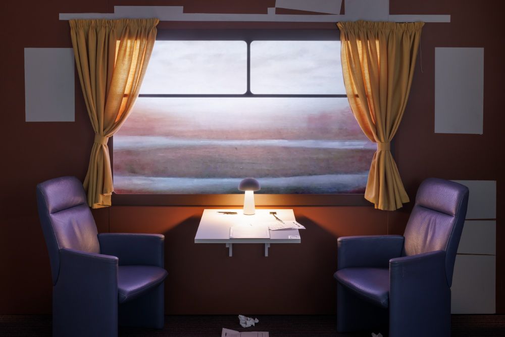 Photo of a small-scale scene inside of a model train. Two empty seats, a table, and a small lamp.