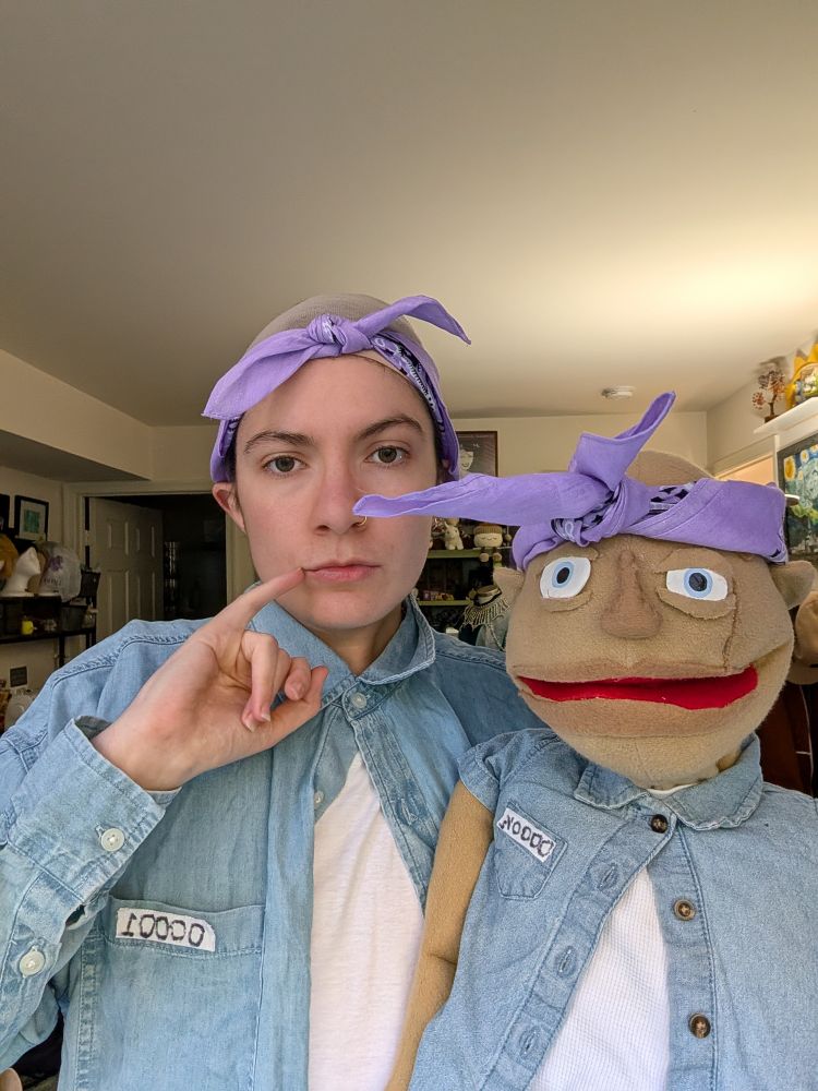 Rio and their mini me puppet dressed as dr evil and mini me in their jail house outfits from Austin powers . They are both in white tshirts with blue denim shirts over them buttoned only at the top.
They both have a purple bandana around their heads and Rio is wearing a bald cap