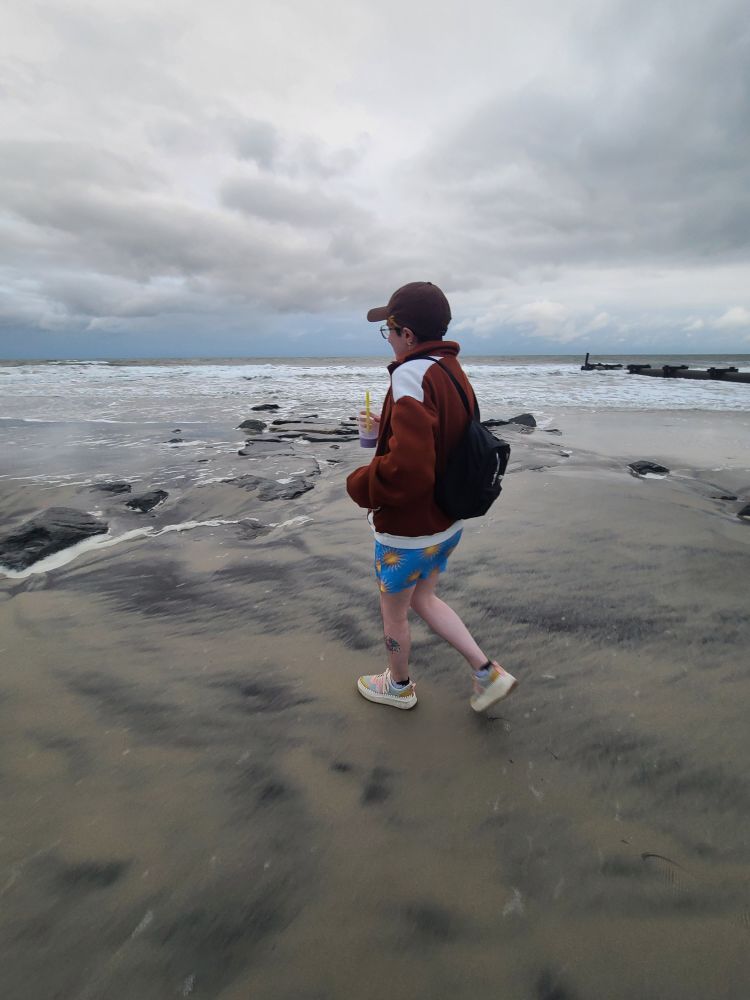 Rio is walking along the beach facing away from the camera, he is holding a boba tea in one hand. A black backpack is on his back and he is wearing a brown hat, a brown zip up hoodie, and blue swim trunks with yellow suns on them.