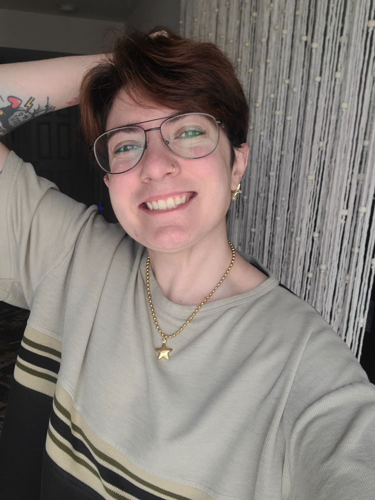 Rio is posing with one hand behind their head and smiling really big. They have reddish brown hair, black aviator glasses, gold jewelry, and a khaki and black striped shirt on