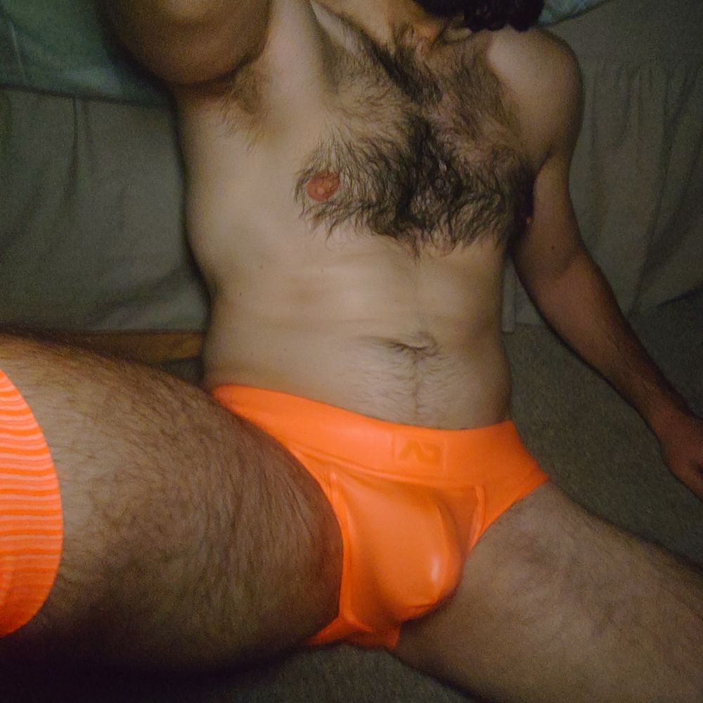 Lowlight selfie of man sitting in tight orange briefs, showing off his hairy chest and legs
