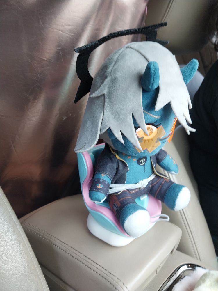 goodsmile happy chaos plushie sitting in a small booster seat (meant for smaller plushies) buckled in. the seatbelt is moments away from popping.