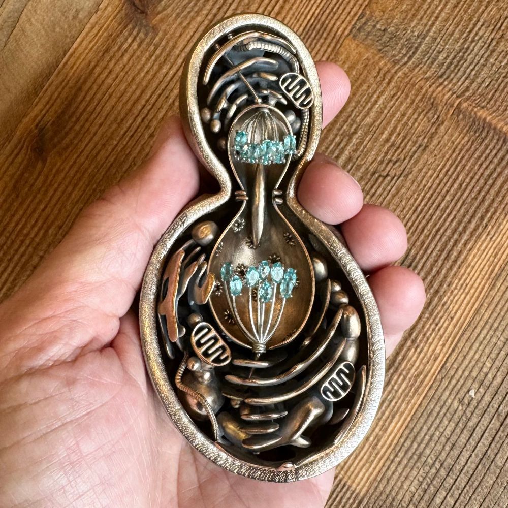 A budding yeast cell in the late anaphase stage of mitosis, a cutaway sculpture in metal. The cell wall is 3D printed steel, the internal organelles are cast bronze, the mitotic spindle is sterling silver, and the chromosomes are apatite gemstones mounted, and silver. Held in my hand over a wooden table.