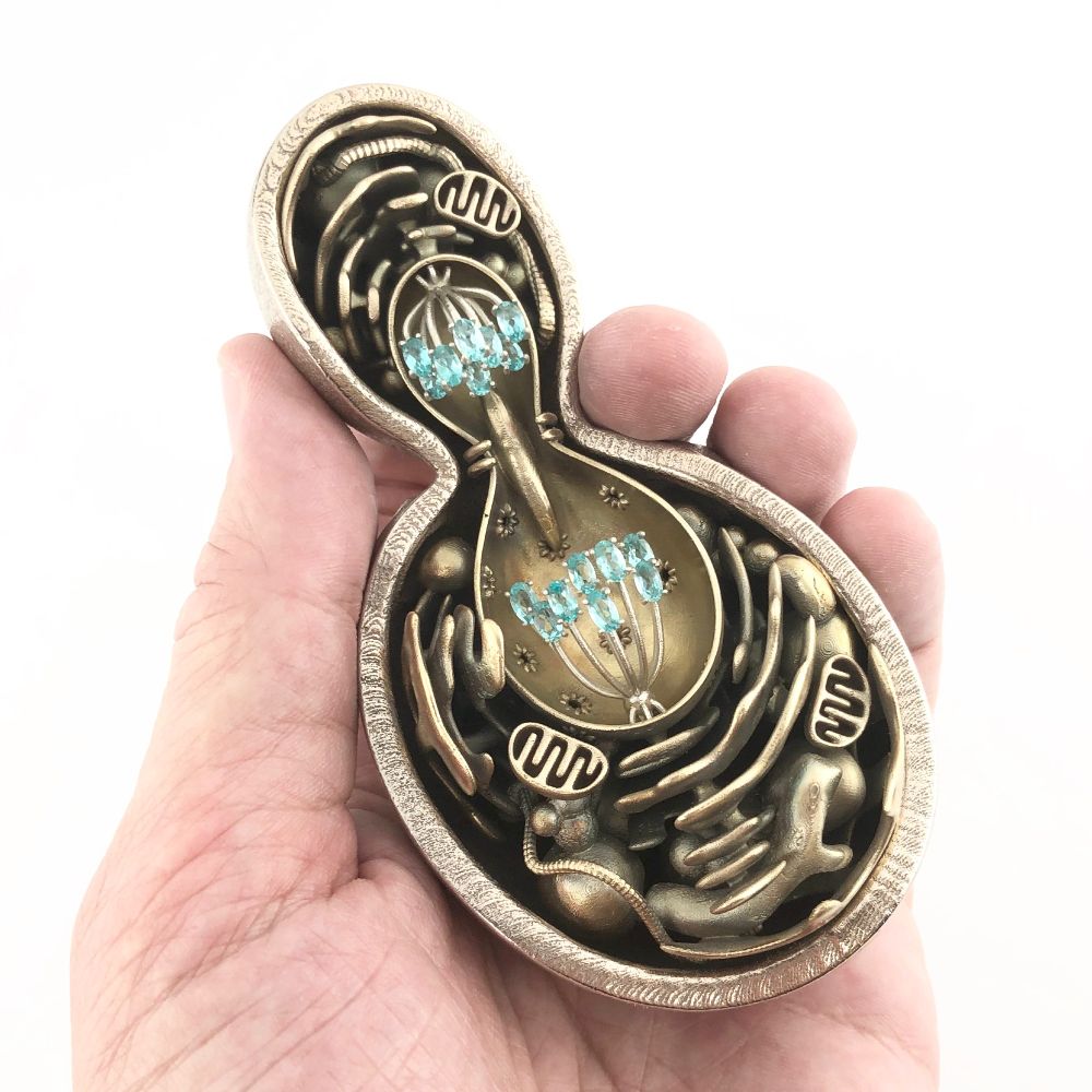 A metal sculpture of a budding yeast cell (Saccharomyces cerevisiae) in late cell division, with a large mother cell budding off a smaller daughter cell, in my hand against a white background. The cell wall is cut away to show many complex, fluid internal structures, including the nucleus and gemstone chromosomes separating after replication, mitochondria with little internal ribbed membranes, the endoplasmic reticulum, and many more. Cell wall is steel, organelles are bronze, mitotic spindle is silver, and the gemstones are apatite, which gets its blue-green color from phosphorus--the element that forms the backbone of DNA.