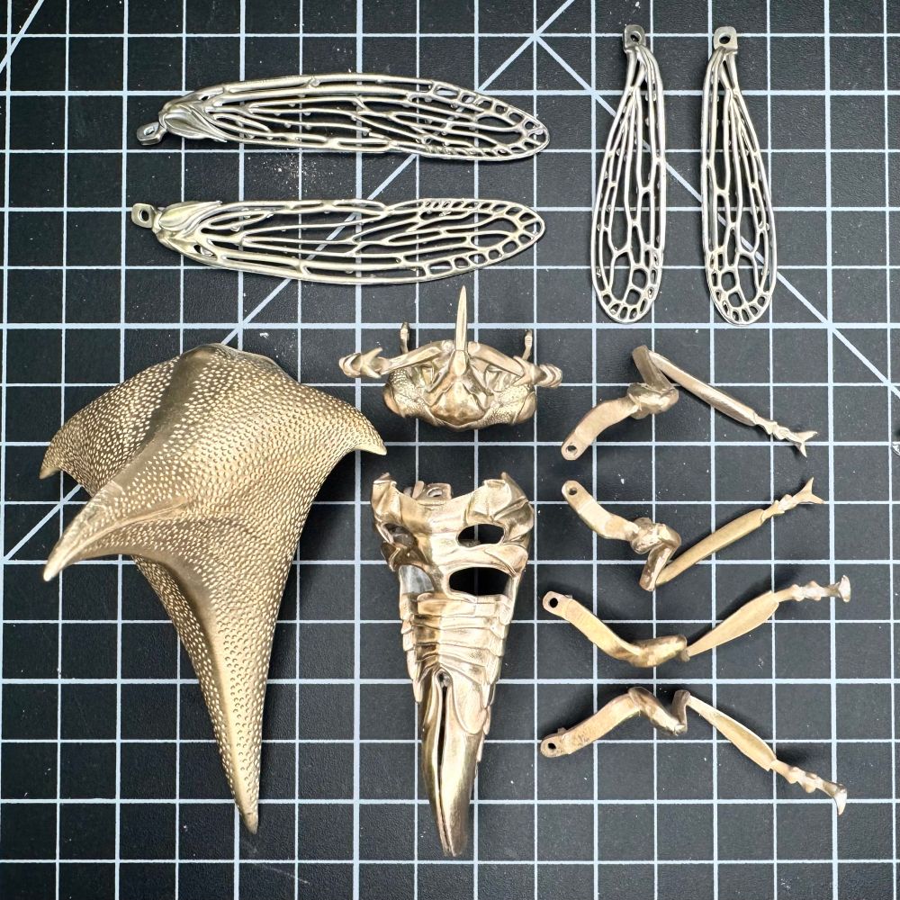 Pieces and parts of a bronze and silver thorn bug laid out on a cutting mat. There are two pairs of silver wings, and seven pieces of the bug.