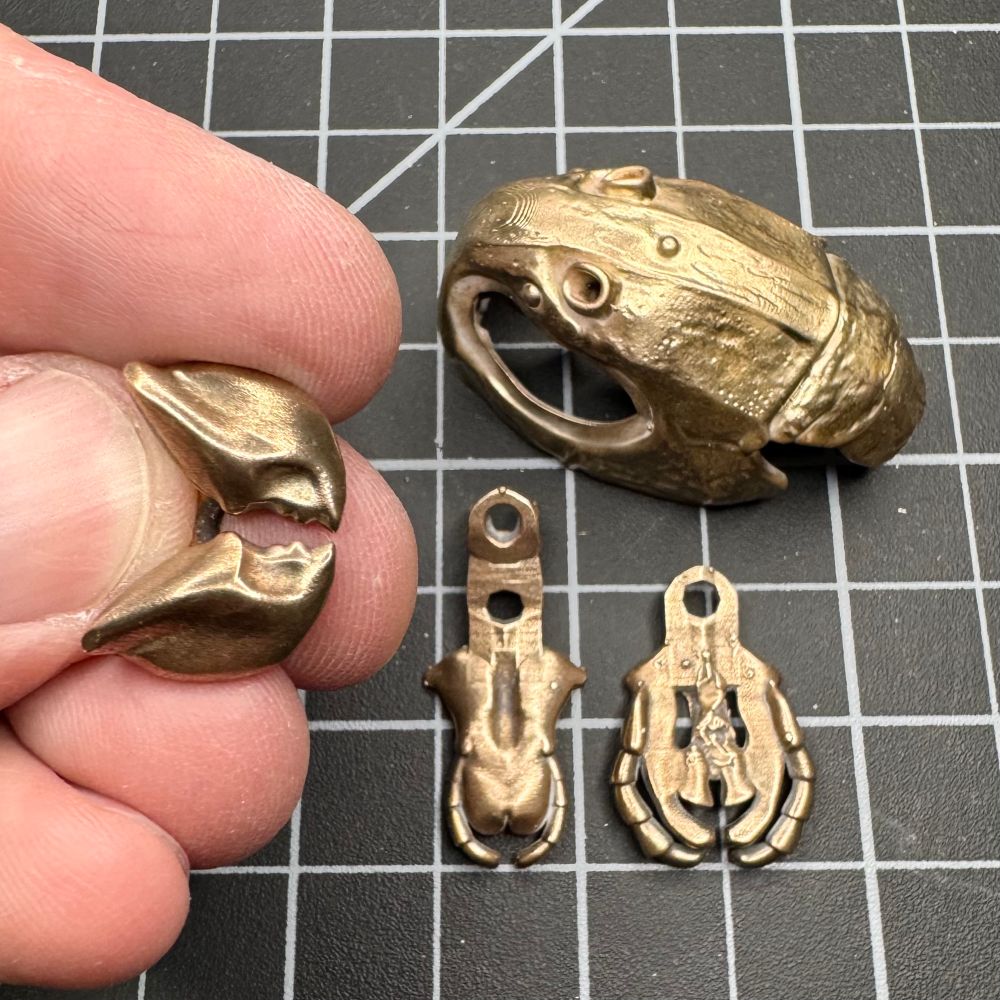 A bronze grasshopper head and three mouth parts on a cutting mat. I’m holding the bronze mandibles in my fingers on the left.