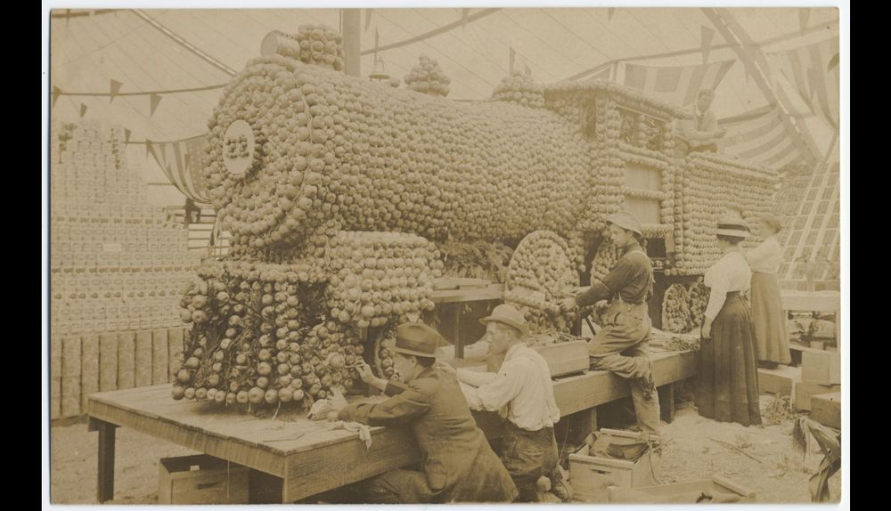 Sepia toned image of 3 men and 2 women working on a life sized model of a train engine made out of apples. 