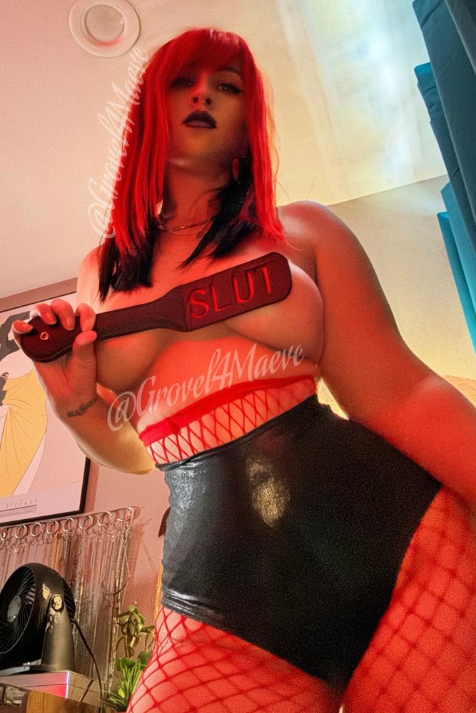 Mistress Maeve with a red wig, red fishnets and high waisted black shorts holding a leather paddle that reads “slut” obstructing her nipples