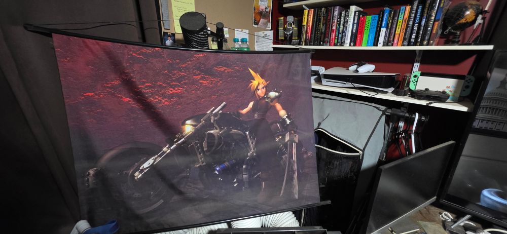 A hanging wall scroll shows Cloud Strife from Final Fantasy VII standing beside the Hardy Daytona motorcycle with his sword, against a textured red background. The wall scroll is displayed in a home office with bookshelves holding books, gaming consoles, and electronics in the background.