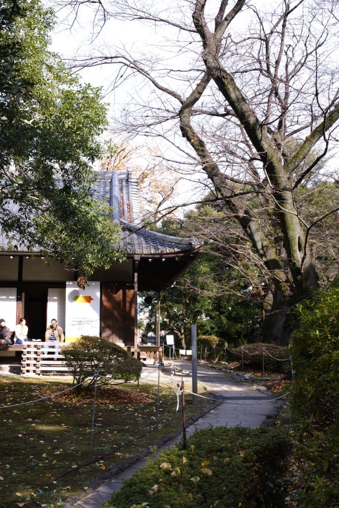 A serene outdoor view of a traditional Japanese structure surrounded by lush greenery. The wooden building has a sloping roof and is set within a well-maintained garden, with pathways lined by low ropes. People sit on the wooden porch, enjoying the peaceful environment.