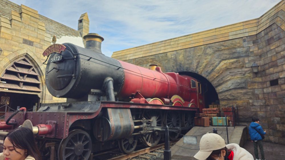 The iconic red locomotive of the Hogwarts Express, marked with "5972" and "Hogwarts Castle," stationed near an arched stone tunnel. Passersby are visible in the foreground, some wearing winter jackets and hats.