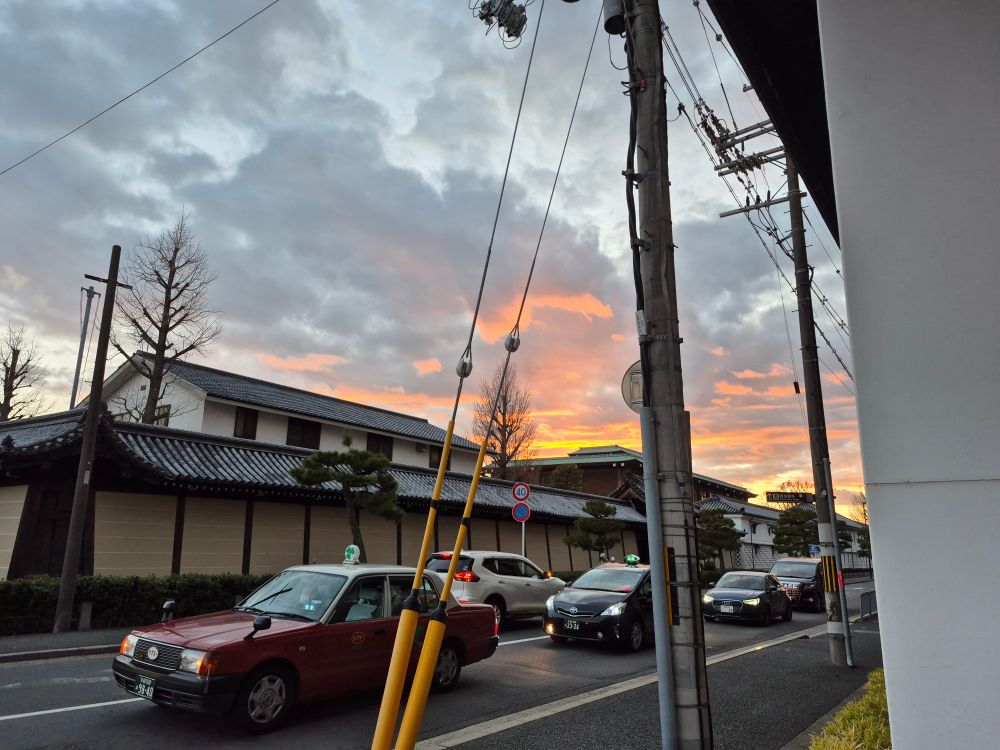 A vibrant urban sunset with orange and pink hues lighting up the clouds. The foreground features traditional Japanese roof tiles, pine trees, and passing cars, alongside utility poles with visible cables, adding a modern touch.