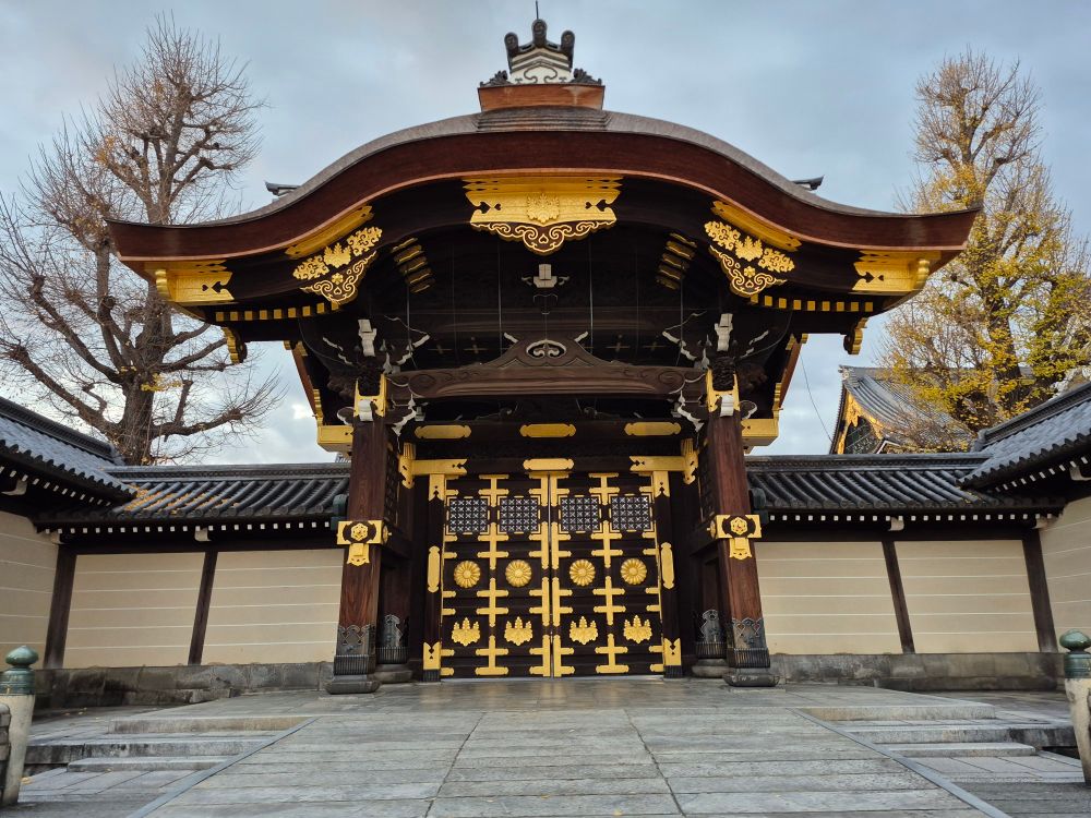 A grand entrance gate of the Imperial Palace in Kyoto, characterized by ornate golden decorations and dark wooden beams. The gate is flanked by traditional white walls with black tiles, set against the backdrop of bare winter trees and a gray sky.