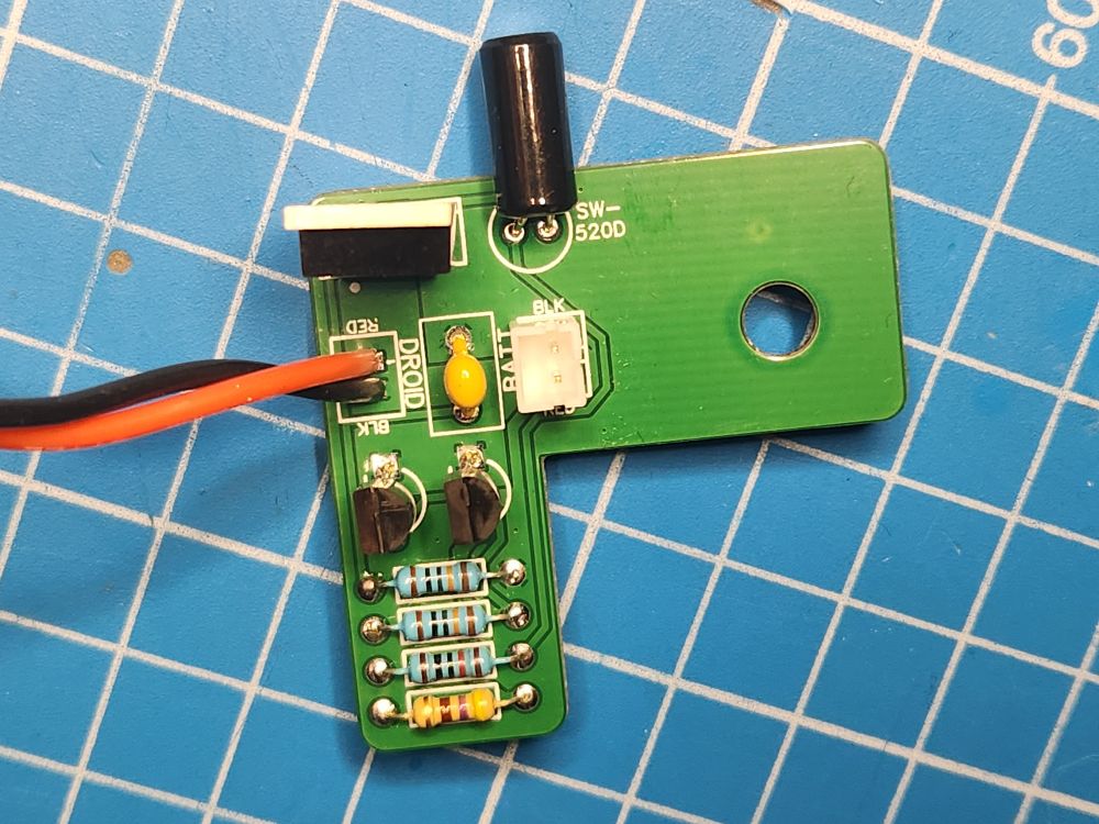The tilt switch mod for new BB droids from Galaxy's Edge. The PCB is L shaped with a hole in it. The idea is to use the hole to align the PCB with the motor housing in BB.

The PCB consists of 4 resistors, 2 2N7000 N-Channel mosfets, a ceramic capacitor, a power transistor, and an SW-520D tilt switch oriented so it sticks up (and is this making a connection) while the BB is in operation.