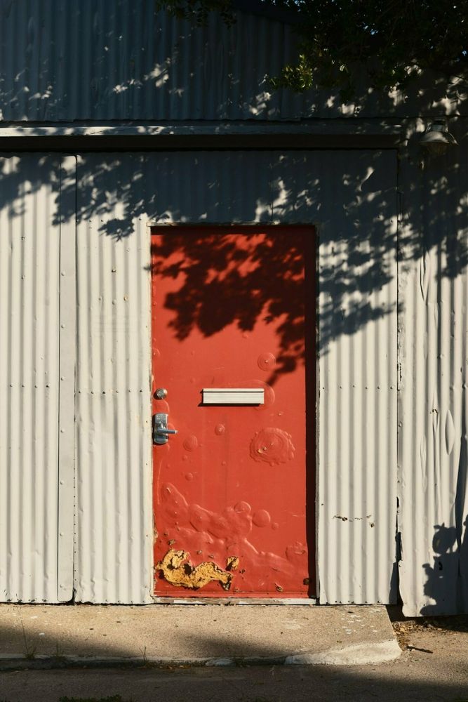Photo of bright red door with paint bubbling off and a section with the inner core of foam visible. The wall around the door is corrugated metal and there is a dark shadow of a tree branch covering the top third of the photo.