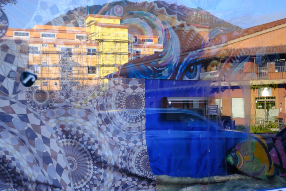 Photo of reflection in a window with a mosaic pattern, a building under construction, a colorful large fish piece of art, another building, an eye, a mountain and a car creating an abstract image.