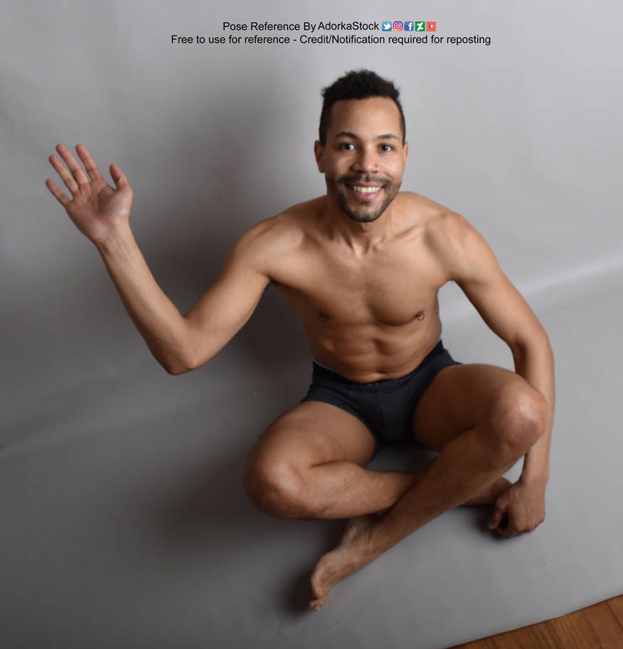 A latino man sitting cross legged on the floor and waving to the camera while smiling.