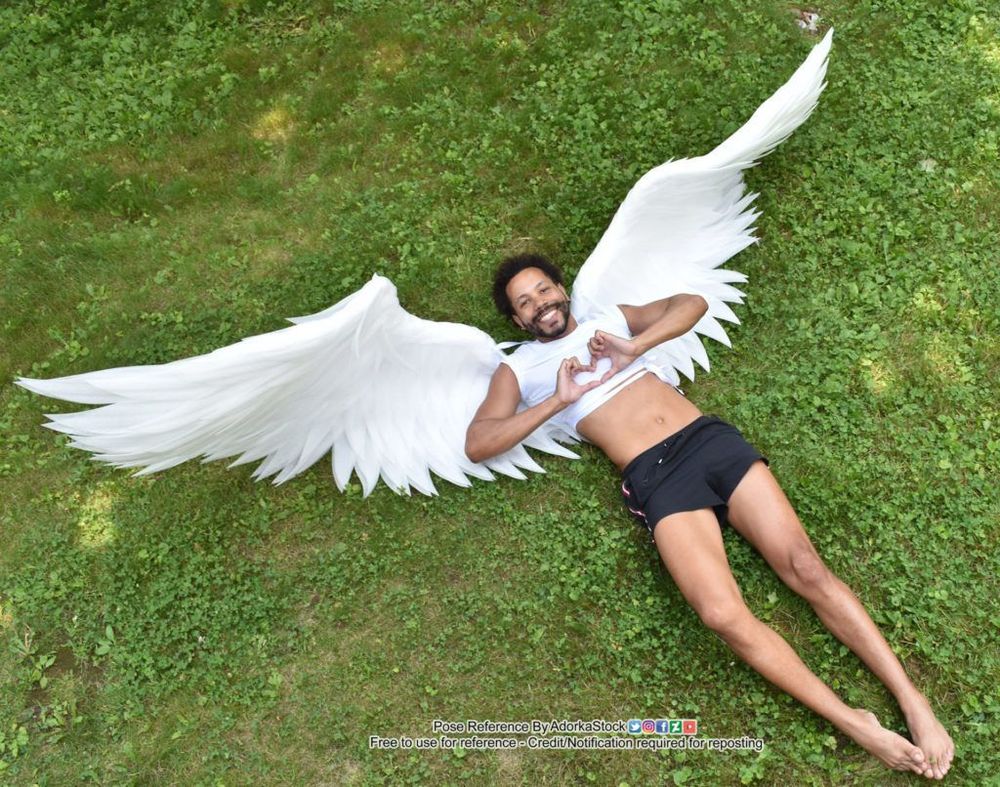 Man wearing large white wings lying on his back making a heart shape with his fingers and smiling.
