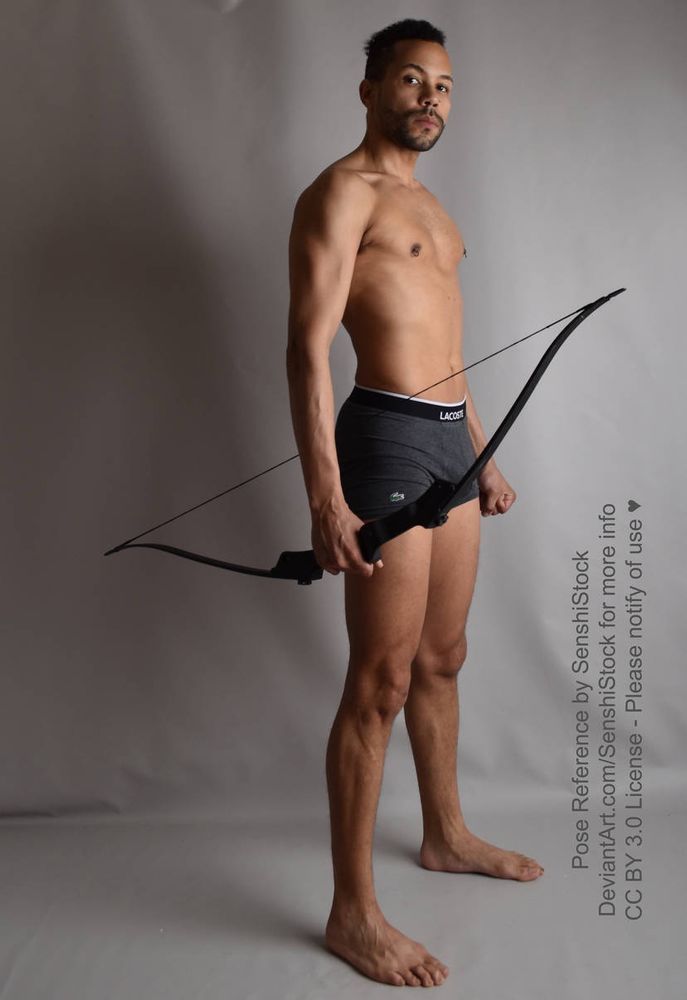 A dark skinned latino man standing in 3/4 low angle view, holding a bow in his right hand, forming a fist with his left, looking at the camera.