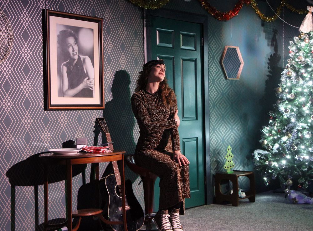 A woman in a long dress is seated in a room with a Christmas tree and a guitar. There is a large black and white portrait of Gloria Swanson on the wall.