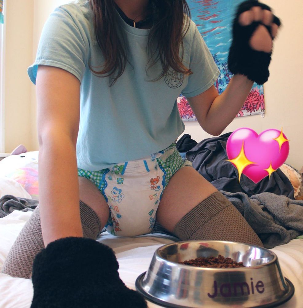 jey, kneeling on her bed next to a dog bowl full of cocoa pebbles :3

she’s wearing brown thigh-highs, her purple collar, a pastel blue tee, a pair of black (kitty >~<) paws, and a well-rounded, soggy little paws diaper 🥰

she’s waving with her left paw and her right is extended on the bed next to the bowl (labeled “jamie”). her wavy brown hair curls over her shoulders.

a shiny heart is applied to pup’s right 💖