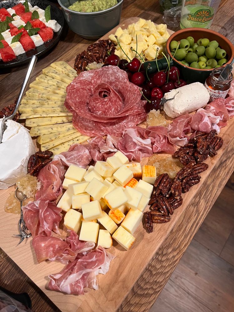 A large wooden serving board, adorned with a spread consisting of a wheel of brie, a river of prosciutto, fanned out triangles of chimichurri Gouda, a pile of cubed Munster, a rose of Italian dry salami, a log of fig and honey goat cheese, a mound of Kerrygold aged cheddar, a bowl of castelvetrano olives, handfuls of candied pecans, bits of honeycomb, cherries, and a tiny jar of Mike’s Hot Honey. Out of frame are two small bowls of fig spread and whole grain mustard. 