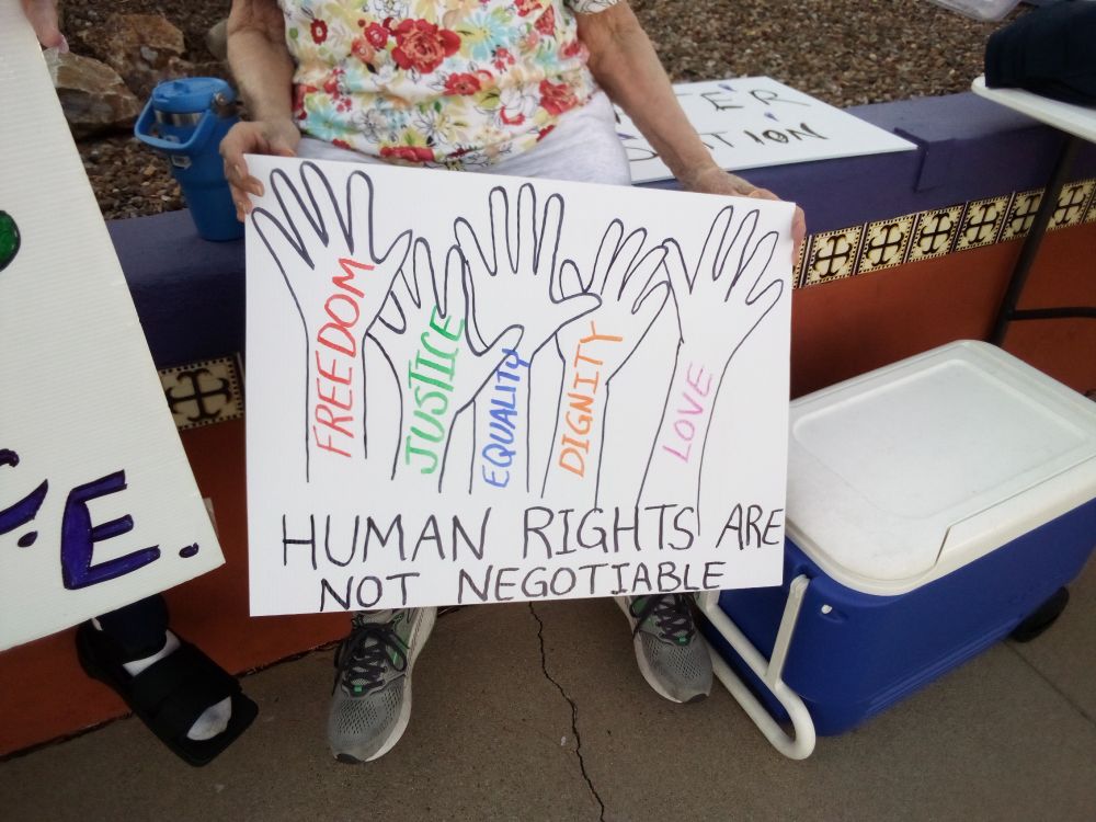Protester holding a sign that reads "Human Rights Are Not Negotiable." The sign has images of outstretched arms with open hands. "Freedom, Justice, Equality, Dignity, and Love" is written on the arms.